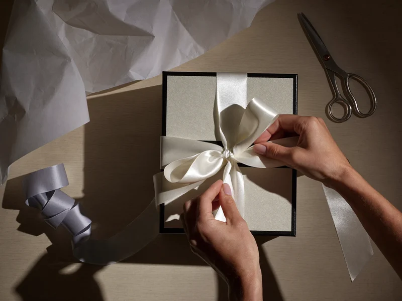 Hände binden eine Schleife um ein eingepacktes Geschenk, daneben Seidenpapier